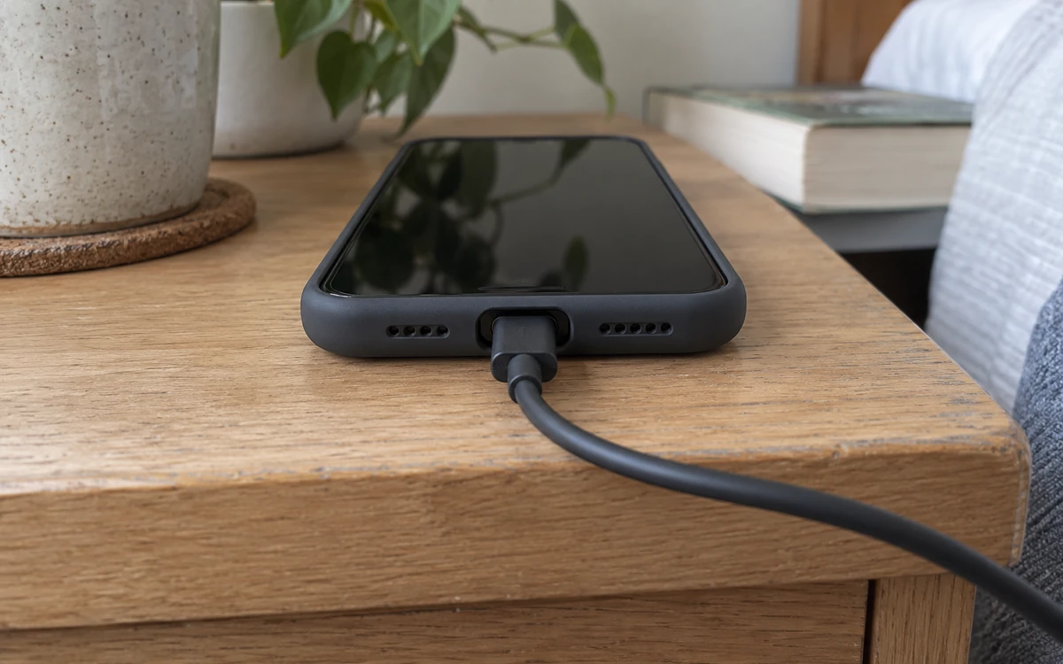 Photo-style illustration of a USB-C cable connected to a cased phone beside an ordinary bedside table, with loose cable slack