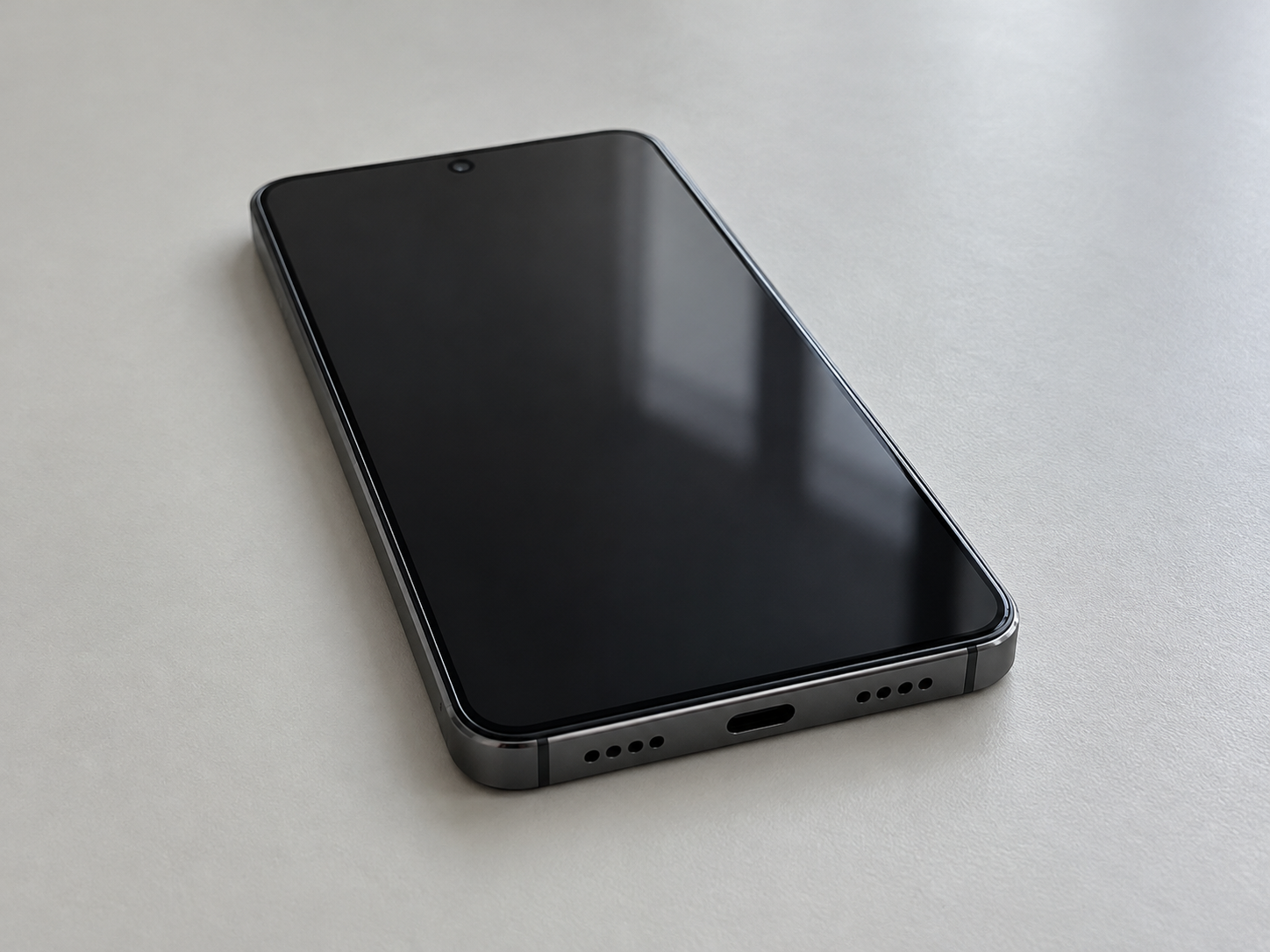An unbranded smartphone lying face up on a plain desk, screen off, with a centered round punch-hole and a USB-C port.